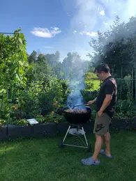 Ein Mann grillt im Freien auf einem Grill, während Rauch aufsteigt. Er steht in einem Garten mit Pflanzen und einem blauen Himmel im Hintergrund. Der Mann trägt eine kurze Hose und Sandalen und scheint konzentriert auf das Grillen zu sein.