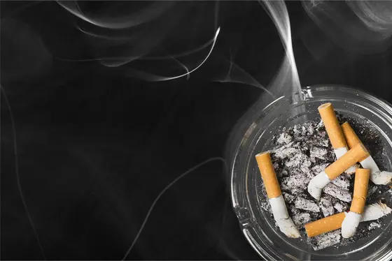 Ein Aschenbecher mit verbrannten Zigarettenstummel und grauem Aschematerial. Dünner Rauch zieht auf und umgibt den Aschenbecher, während der Hintergrund schwarz ist.