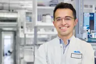 Ein junger Wissenschaftler in einem weißen Laborkittel lächelt freundlich. Er steht in einem modernen Labormilieu mit Regalen voller wissenschaftlicher Geräte im Hintergrund. Sein Name und die Institution sind auf einem Namensschild sichtbar.