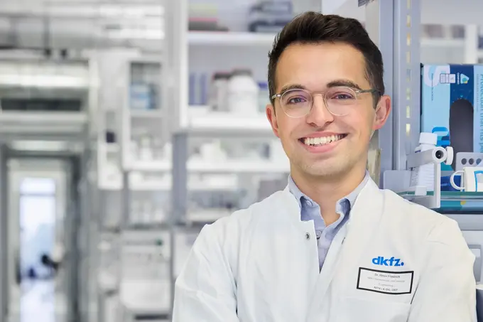 Ein junger Wissenschaftler in einem weißen Laborkittel lächelt freundlich. Er steht in einem modernen Labormilieu mit Regalen voller wissenschaftlicher Geräte im Hintergrund. Sein Name und die Institution sind auf einem Namensschild sichtbar.