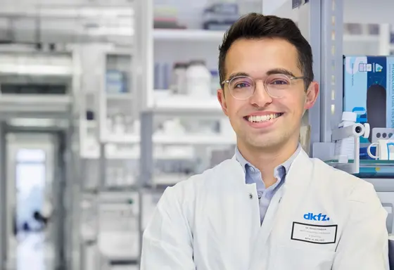 Ein junger Wissenschaftler in einem weißen Laborkittel lächelt freundlich. Er steht in einem modernen Labormilieu mit Regalen voller wissenschaftlicher Geräte im Hintergrund. Sein Name und die Institution sind auf einem Namensschild sichtbar.