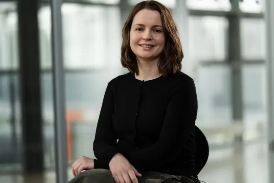 A woman with shoulder-length brown hair smiles while sitting on a chair in an indoor space. She wears a black cardigan and appears to be engaged and approachable, reflecting positivity and confidence. The background features large windows and a modern interior design.