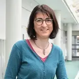 A woman with shoulder-length brown hair and glasses stands next to a railing, smiling. She is wearing a teal sweater over a pink shirt. The background features a modern, well-lit interior space.