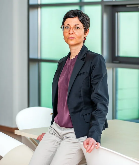 Eine Frau sitzt auf einem Tisch in einem modernen, hellen Büro. Sie trägt einen schwarzen Blazer und ein lila Oberteil. Im Hintergrund sind große Fenster sichtbar, die Tageslicht hereinlassen.