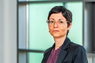 Eine Frau sitzt auf einem Tisch in einem modernen, hellen Büro. Sie trägt einen schwarzen Blazer und ein lila Oberteil. Im Hintergrund sind große Fenster sichtbar, die Tageslicht hereinlassen.