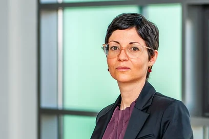 Eine Frau sitzt auf einem Tisch in einem modernen, hellen Büro. Sie trägt einen schwarzen Blazer und ein lila Oberteil. Im Hintergrund sind große Fenster sichtbar, die Tageslicht hereinlassen.