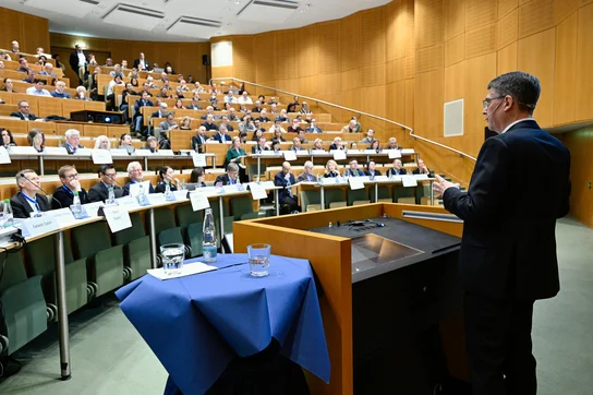 Ein Redner steht hinter einem Pult und spricht zu einem Publikum in einem großen Saal. Die Zuschauer sind in Reihen angeordnet, viele von ihnen haben Namensschilder vor sich. Der Raum ist hell erleuchtet und hat eine moderne Holzverkleidung.