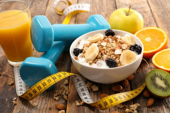 Ein gesundes Frühstück mit einer Schüssel Müsli, belegt mit Bananen, Heidelbeeren und Nüssen. Neben der Schüssel steht ein Glas Orangensaft und Fitness-Hanteln. Um die Szene ist ein Maßband gelegt, zusammen mit einer grünen Kiwi, einem Apfel und einer Orange.