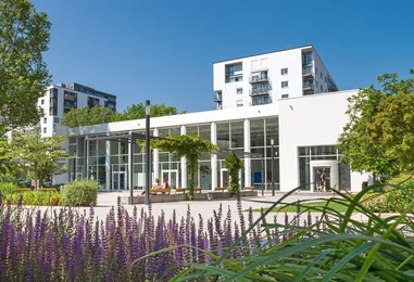 Eine moderne Gebäudeansicht mit großen Fenstern und einem Außenbereich. Im Vordergrund blühen lila Pflanzen, während im Hintergrund moderne Wohngebäude zu sehen sind. Menschen sitzen entspannt auf Bänken im Freien. Der Himmel ist klar und blau.