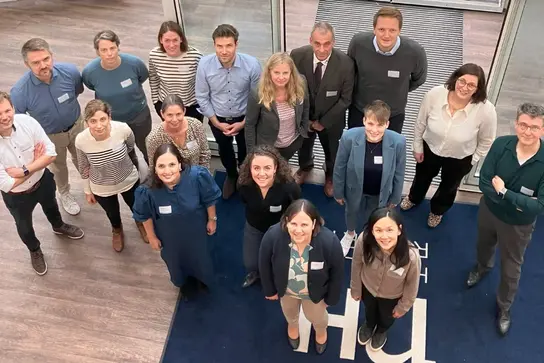 Gruppe von 16 Personen steht in einem Eingangsbereich und blickt nach oben in die Kamera. Die Personen tragen Namensschilder und sind in Business-Casual-Kleidung gekleidet.