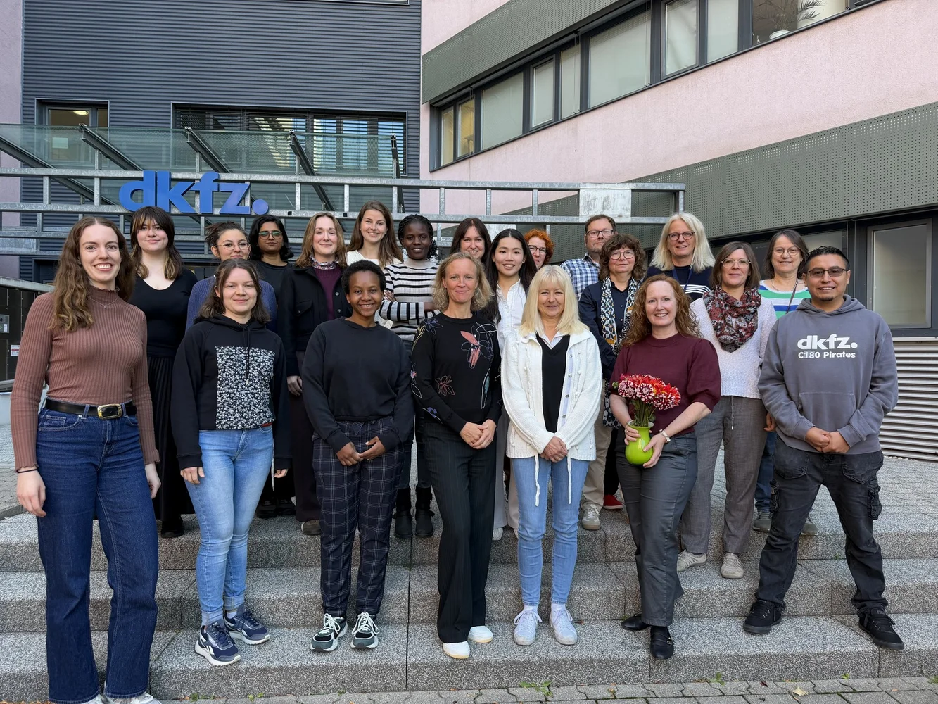 Eine Gruppe von etwa 20 Personen steht auf den Treppen vor einem Gebäude mit dem Logo "dkfz". Die Teilnehmer bestehen aus Frauen und Männern unterschiedlichen Alters und ethnischer Herkunft. Einige Personen halten Blumen oder Töpfe. Es herrscht eine freundliche, gemeinschaftliche Atmosphäre.