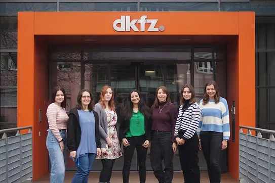 Eine Gruppe von acht Frauen steht vor dem Eingang des DKFZ (Deutsches Krebsforschungszentrum). Sie lächeln und tragen verschiedene Kleidung. Der Hintergrund zeigt eine moderne Gebäude-Architektur mit einem orangefarbenen Schriftzug "dkfz".