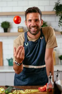 Mann in Küchenschürze wirft lächelnd eine Tomate in die Luft; Gemüse liegt auf der Arbeitsfläche.
