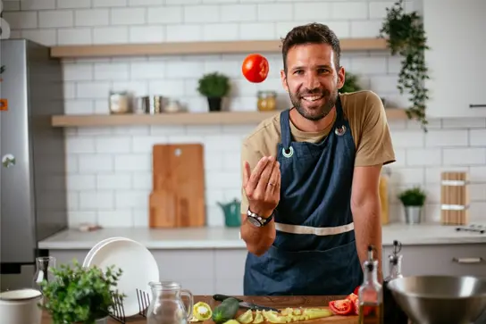 Mann in Küchenschürze wirft lächelnd eine Tomate in die Luft; Gemüse liegt auf der Arbeitsfläche.