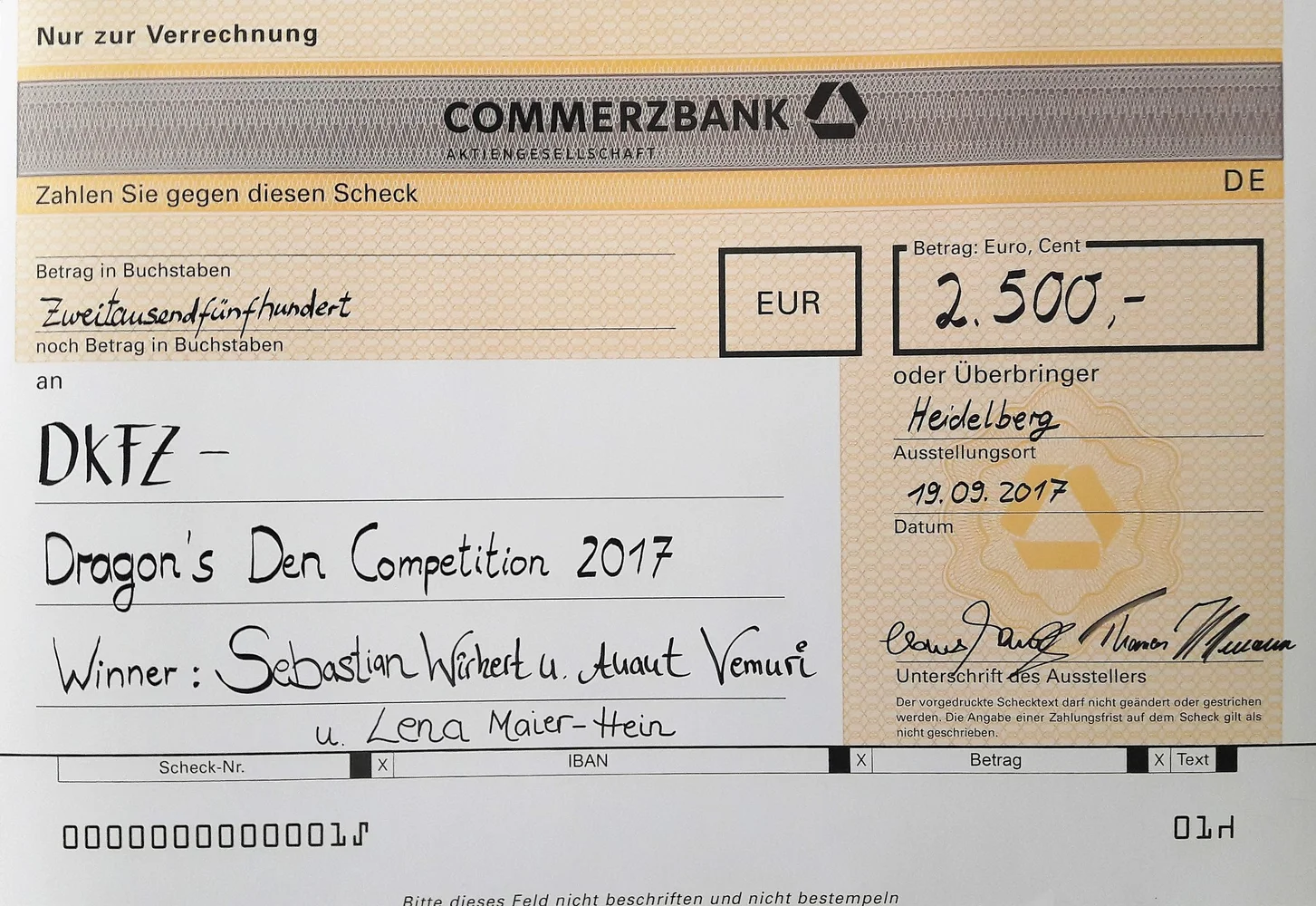 Alternative text: "A large bank check from Commerzbank for €2,500, awarded in 2017 for the DKFZ Dragon's Den Competition. The check is presented to winners Sebastian Wichert, Ahmad Vemuri, and Lena Maur-Hein, dated September 19, 2017, with Heidelberg as the place of issue."