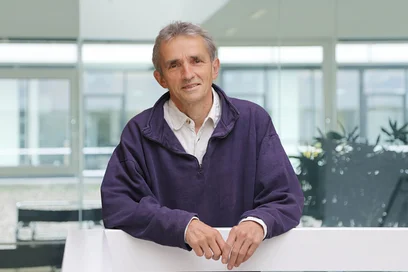 Ein Mann steht an einem Geländer in einem modernen Raum mit großen Fenstern. Er trägt einen lila Pullover über einem weißen Hemd und lächelt. Im Hintergrund sind Pflanzen und Möbel zu sehen.