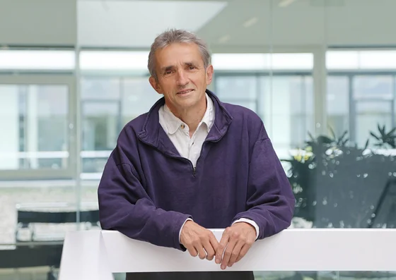 Ein Mann steht an einem Geländer in einem modernen Raum mit großen Fenstern. Er trägt einen lila Pullover über einem weißen Hemd und lächelt. Im Hintergrund sind Pflanzen und Möbel zu sehen.