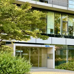 Das Bild zeigt das Eingangstor des DKFZ (Deutsches Krebsforschungszentrum) umgeben von Grünanlagen. Die moderne Architektur beinhaltet große Fenster, durch die viel Licht fällt. Der Name "dkfz" ist deutlich am Eingang zu erkennen.