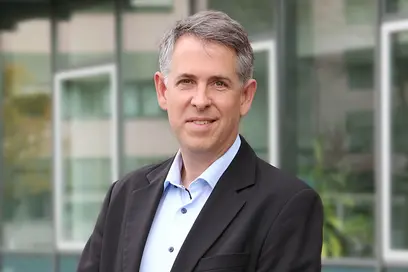 Ein Mann steht vor einem modernen Gebäude. Er trägt ein schwarzes Jackett und ein hellblaues Hemd. Der Gesichtsausdruck ist freundlich, während er in die Kamera lächelt. Im Hintergrund sind Fenster und Pflanzen zu sehen.