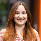 Eine Frau mit langen, rotbraunen Haaren lächelt in die Kamera. Sie trägt ein shirt mit einem bunten, geometrischen Muster. Der Hintergrund zeigt unscharfe grüne und orangefarbene Elemente, die auf eine Außenumgebung hindeuten.