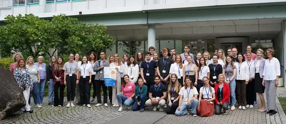 Eine große Gruppe von Menschen steht vor einem modernen Gebäude. Die Teilnehmer sind in unterschiedlichen Kleidungsstilen gekleidet und versammeln sich lächelnd für ein Gruppenfoto. Im Hintergrund sind Bäume und Fahrräder sichtbar, die zur Umgebung beitragen.