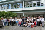 Eine große Gruppe von Menschen steht vor einem modernen Gebäude. Die Teilnehmer sind in unterschiedlichen Kleidungsstilen gekleidet und versammeln sich lächelnd für ein Gruppenfoto. Im Hintergrund sind Bäume und Fahrräder sichtbar, die zur Umgebung beitragen.