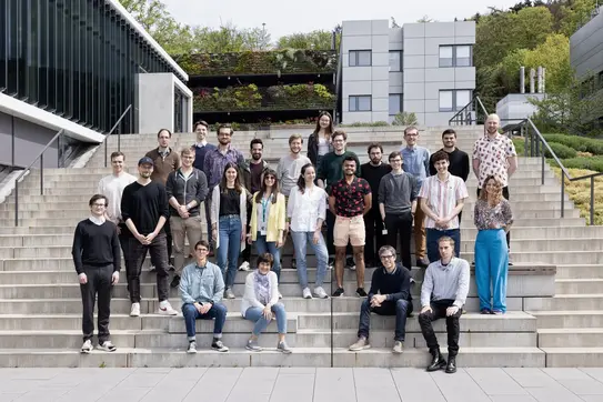 Eine Gruppe von etwa 30 Personen steht auf einer breiten Treppe vor modernen Gebäuden und einer grünen Landschaft. Die Mitglieder sind in verschiedenen Kleidungsstilen gekleidet und lächeln, während sie für das Gruppenfoto posieren.