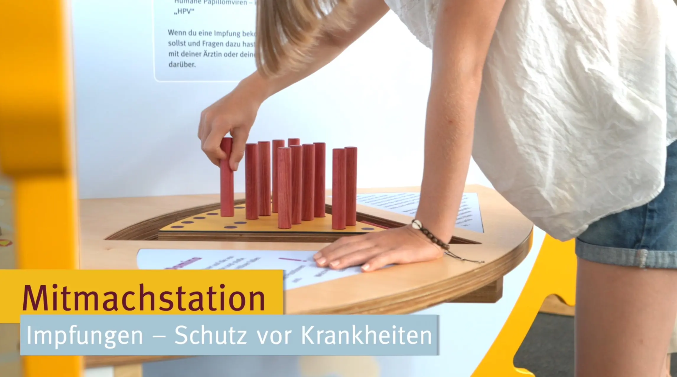 Die Abbildung zeigt eine Mitmachstation, an der eine Person mit roten Stäben interagiert. Der Fokus liegt auf dem Thema Impfungen und deren Schutz vor Krankheiten, was durch die stationäre Gestaltung unterstützt wird.