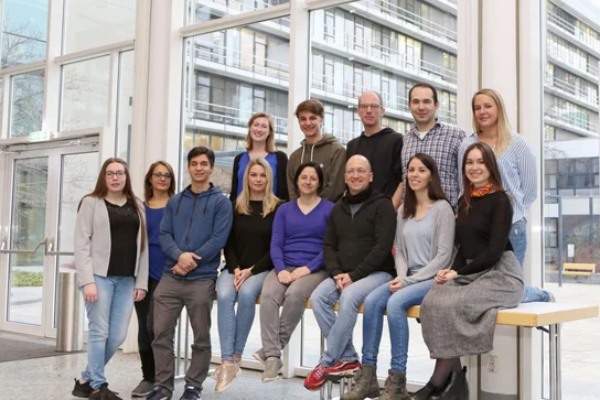 A diverse group of fifteen individuals is gathered in a well-lit indoor space with large windows. They are standing and sitting in two rows, smiling at the camera. The setting appears modern and is likely an educational or professional environment.