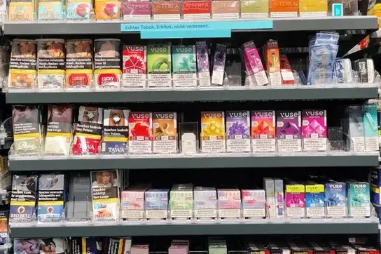 Ein Regal in einem Geschäft für Tabakwaren mit verschiedenen Sorten von Zigaretten und Dampferprodukten. Mehrere Marken und Geschmacksrichtungen sind klar aufgereiht, darunter auch Hinweise zu den gesundheitlichen Risiken des Rauchens. Das obere Schild zeigt "Tabakasse".