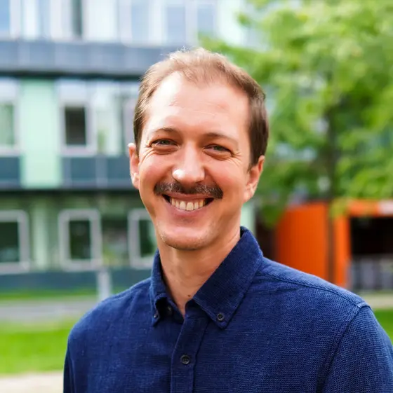 Ein lächelnder Mann steht im Freien vor einem modernen Gebäude mit großen Fenstern. Er trägt ein blaues Hemd und hat einen Schnurrbart. Die Umgebung ist grün mit Bäumen und Rasenflächen, was ein freundliches und einladendes Ambiente schafft.