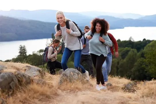 Gruppe junger Menschen wandert lachend bergauf mit Blick auf See und Wald.