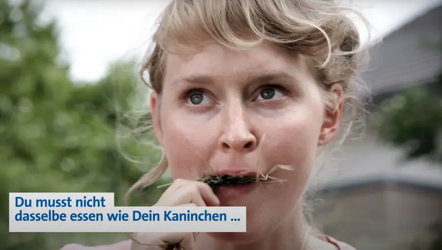 Frau kaut an Grashalmen, Blick nach vorn; eingeblendeter Text: „Du musst nicht dasselbe essen wie Dein Kaninchen …“
