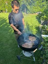 Ein Mann grillt in einem Garten, während Rauch über den Grill aufsteigt. Er trägt Sonnenbrillen und schaut konzentriert auf das Fleisch, das auf dem Grill zubereitet wird. Im Hintergrund sind grüne Pflanzen und Rasen zu sehen.