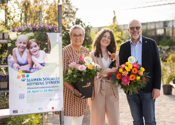 Drei Personen stehen in einem Garten und halten bunte Blumensträußchen. Hinter ihnen ist ein Plakat mit dem Motto „Blumen schenken, Hoffnung spenden“ zu sehen. Die Aktion zielt darauf ab, Spenden für die Unterstützung von krebskranken Kindern zu sammeln und läuft vom 28. April bis 24. Mai 2025.