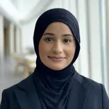 Eine junge Frau mit dunklem Hijab und formeller Jacke lächelt freundlich in die Kamera. Der Hintergrund zeigt einen modernen Innenraum mit hellen Farben.