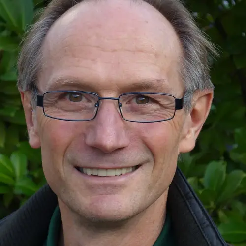 A middle-aged man with short hair and glasses smiles warmly, wearing a dark jacket over a green shirt. He is set against a background of green foliage, suggesting an outdoor setting.