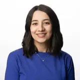 Porträt einer lächelnden Frau mit langen, dunklen Haaren, die einen blauen Pullover trägt. Der Hintergrund ist neutral und hell, was den Fokus auf ihr Gesicht und ihren freundlichen Ausdruck lenkt. Sie wirkt offen und sympathisch.