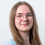 Ein junger Mensch mit langen, blonden Haaren und einer blauen T-Shirt steht vor einem hellen Hintergrund. Sie trägt runde, transparente Brillen und lächelt freundlich.