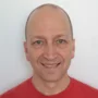 A smiling man with a shaved head stands against a plain background, wearing a red short-sleeve shirt. His hands are clasped behind his back, and he has a friendly expression.