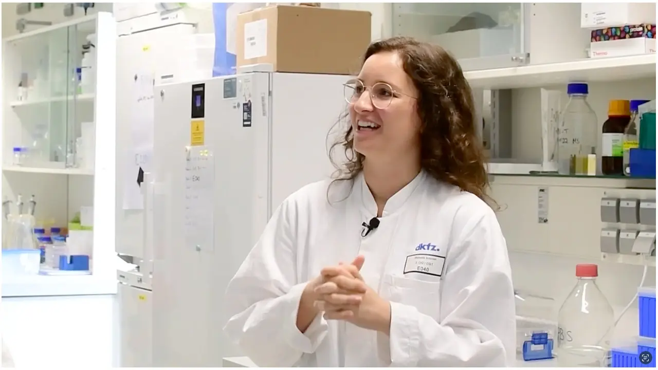 A smiling female scientist in a lab coat stands in a laboratory, engaging with the camera. She has curly hair and glasses. The background includes shelves with laboratory equipment and containers, emphasizing a scientific environment. Her expression conveys enthusiasm and warmth.
