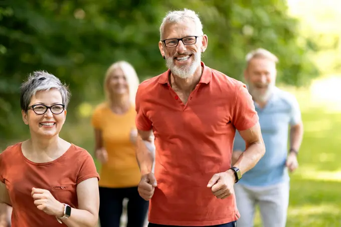 Eine Gruppe von vier Personen, darunter Männer und Frauen mittleren Alters, joggen fröhlich in einer grünen Parklandschaft. Sie tragen sportliche Kleidung und wirken aktiv und gesund. Die Atmosphäre ist positiv und einladend.