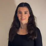 Eine junge Frau mit langen, lockigen, dunklen Haaren steht vor einem hellen Hintergrund. Sie trägt ein schwarzes Oberteil und hat einen freundlichen Gesichtsausdruck.