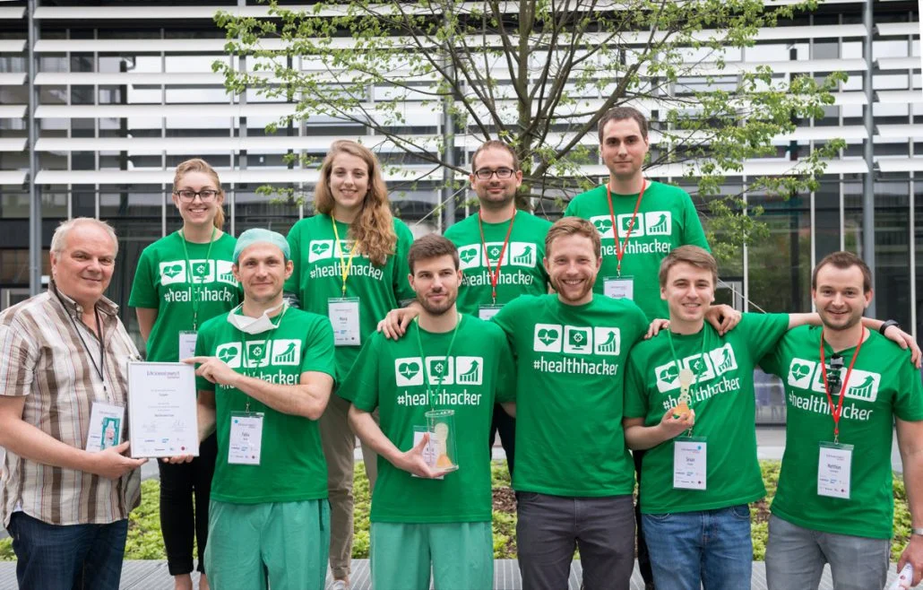 A group of eleven people poses for a photo, wearing green shirts with the text “healthhacker.” They are celebrating an achievement, with some holding trophies and certificates. The background features greenery and a modern building structure.
