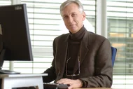 Ein Mann sitzt an einem Schreibtisch vor einem Computer. Er trägt einen dunklen Pullover und einen braunen Blazer. Sein Blick ist freundlich. Im Hintergrund sind Fenster mit horizontalen Jalousien zu sehen.