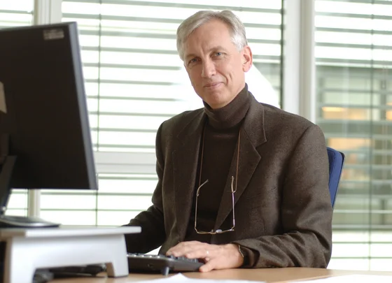 Ein Mann sitzt an einem Schreibtisch vor einem Computer. Er trägt einen dunklen Pullover und einen braunen Blazer. Sein Blick ist freundlich. Im Hintergrund sind Fenster mit horizontalen Jalousien zu sehen.