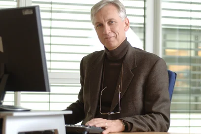 Ein Mann sitzt an einem Schreibtisch vor einem Computer. Er trägt einen dunklen Pullover und einen braunen Blazer. Sein Blick ist freundlich. Im Hintergrund sind Fenster mit horizontalen Jalousien zu sehen.