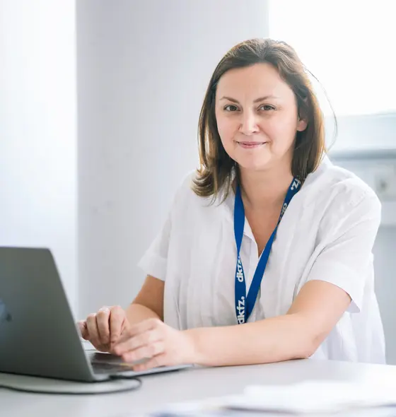 Ana Banito sitzt an einem Tisch mit einem Laptop und lächelt in die Kamera. Sie trägt ein helles, lockeres Hemd und eine ID-Kette. Der Hintergrund zeigt einen hellen und modernen Raum, der für Forschungsarbeiten genutzt wird, passend zu ihrer Rolle als Leiterin einer Nachwuchsgruppe im Bereich Weichteilsarkome.