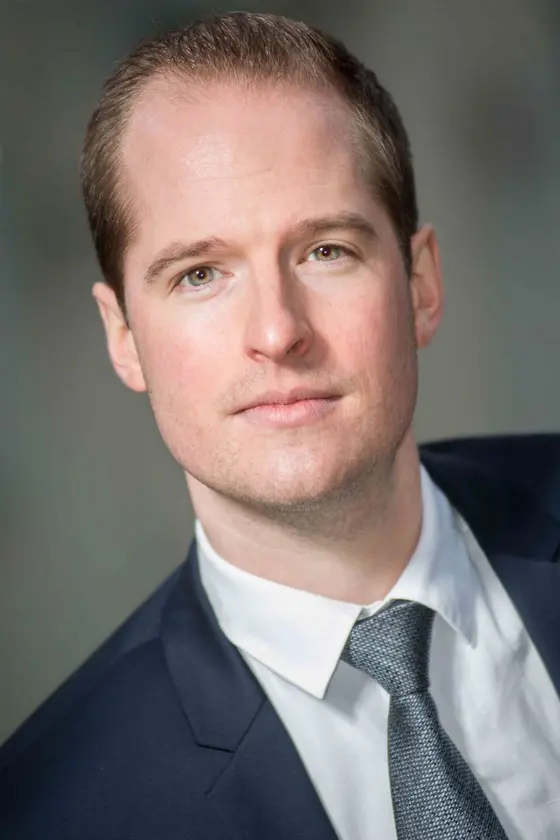 A professional young man is depicted with a slight smile. He has short, light brown hair and is wearing a dark suit with a white shirt and a gray tie. The background is neutral, focusing on his confident expression.