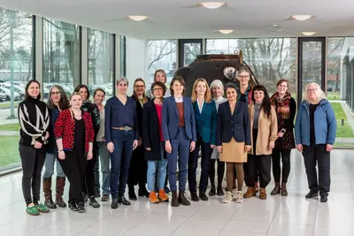Gruppe von 17 Frauen in einem modernen Gebäude, die lächeln und für ein Foto posieren. Sie tragen unterschiedliche Kleidung von casual bis business. Im Hintergrund sind große Fenster sichtbar, durch die Tageslicht strömt, und ein Objekt, das wie ein altes Technikgerät aussieht.